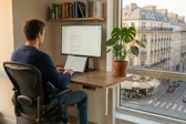 A generated, photorealistic image of a developer in his workspace with a monstera plant, books, and a view of the city of Paris. This could well be me, but some small details don't lie.